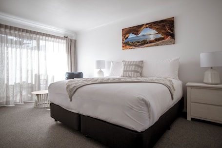 A hotel room with a bed, a nightstand, a lamp, a chair, and a large window with curtains.