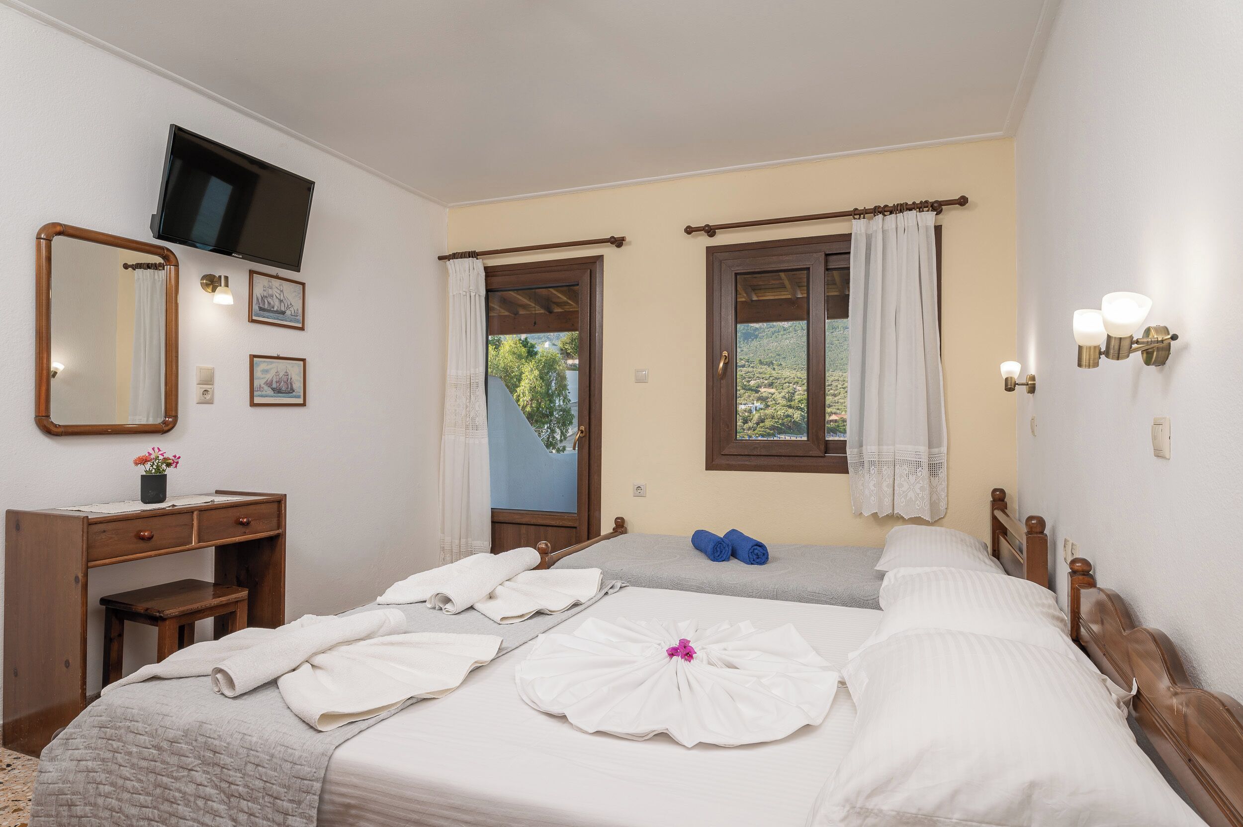A bedroom with two beds, a desk, a mirror, a TV, and a window with curtains.