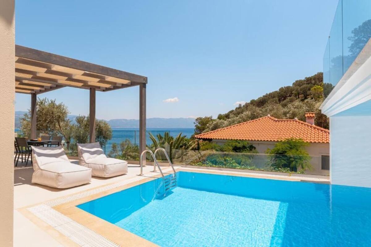 A swimming pool with lounge chairs and a pergola, overlooking a coastal landscape with a view of the sea and hills.