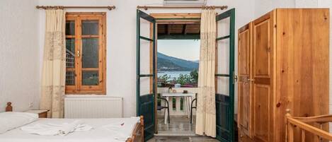 A bedroom with a bed, a wooden wardrobe, and an open door leading to a balcony with a table and chairs.