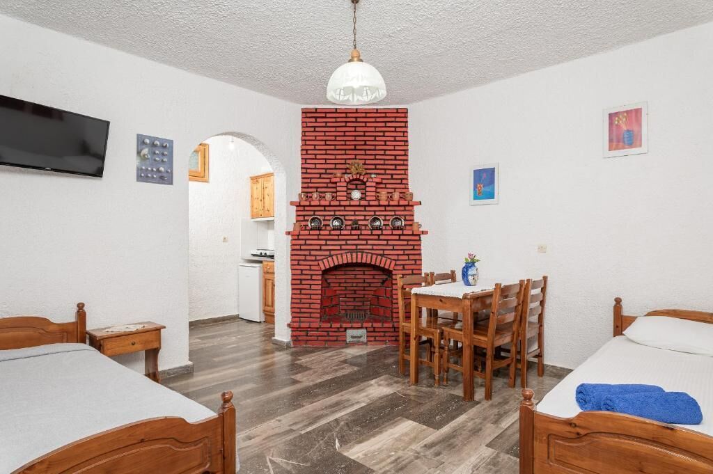 A room with two wooden beds, a brick fireplace, a dining table with chairs, a TV mounted on the wall, and a kitchen area visible through an archway.
