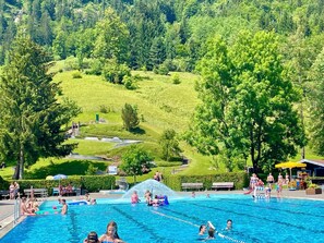 A swimming pool with clear blue water, surrounded by lush green trees and a hilly landscape in the background. - Dosbach Vacation Apartment (Reit im Winkl)
