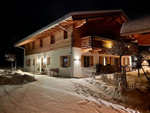 Un chalet en bois à deux étages, doté d’un balcon, avec un sol enneigé et des sièges à l’extérieur.