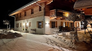 Eine zweistöckige Holzhütte mit Balkon, verschneitem Gelände und einer Bank davor.