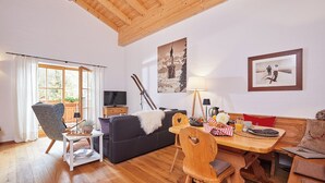 Ein gemütliches Wohnzimmer mit Holzbalkendecke, einer Couch, einem Esstisch mit Stühlen, einem Fernseher und Skiausrüstung.