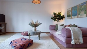 Ein modernes Wohnzimmer mit einem rosafarbenen Sofa, einem runden Couchtisch mit einer Vase getrockneter Pflanzen, einer großen Topfpflanze und einem Fernseher auf einem Holzständer.