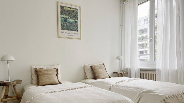 Un dormitorio con dos camas, ropa de cama blanca, un cuadro en la pared y vistas a edificios a través de la ventana.