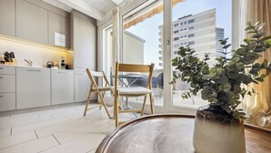 Cozinha moderna com armários brancos, área de jantar com mesa e cadeiras de madeira, e uma janela grande com vista para edifícios.