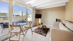 Sala de estar moderna com sofá, cadeiras, mesa de centro e TV. Janelas amplas oferecem vista para a cidade.