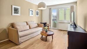 Ein Wohnzimmer mit einem beige farbenen Sofa, einem runden Couchtisch mit einer Vase, einem schwarzen Sessel und einem Fernseher auf einem schwarzen Schrank.