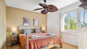 Ein Schlafzimmer mit einem Bett, zwei Nachttischen, einem Deckenventilator und einem Fenster mit Blick auf Bäume.