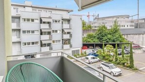 Un balcon avec une chaise verte, une vue sur un parking et, au loin, un immeuble à plusieurs étages.