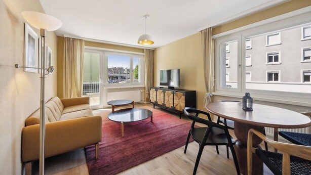 A modern living room with a sofa, a coffee table, a dining table with chairs, a TV on a cabinet, and large windows with a city view. - Luxury Apartment close to the city center! (Zürich)