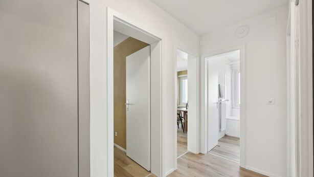 Um corredor com paredes brancas e soalho de madeira, que leva a um quarto com janela e porta.