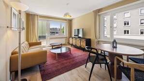 Uma sala de estar moderna com sofá, mesa de centro, mesa de jantar e cadeiras. Há uma televisão em um armário, um abajur e vista de edifícios por amplas janelas.