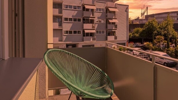 A balcony with a green chair, a view of a multi-story building, and a sunset. - Elegant City Apartment with Balcony! (101-16) (Zürich)