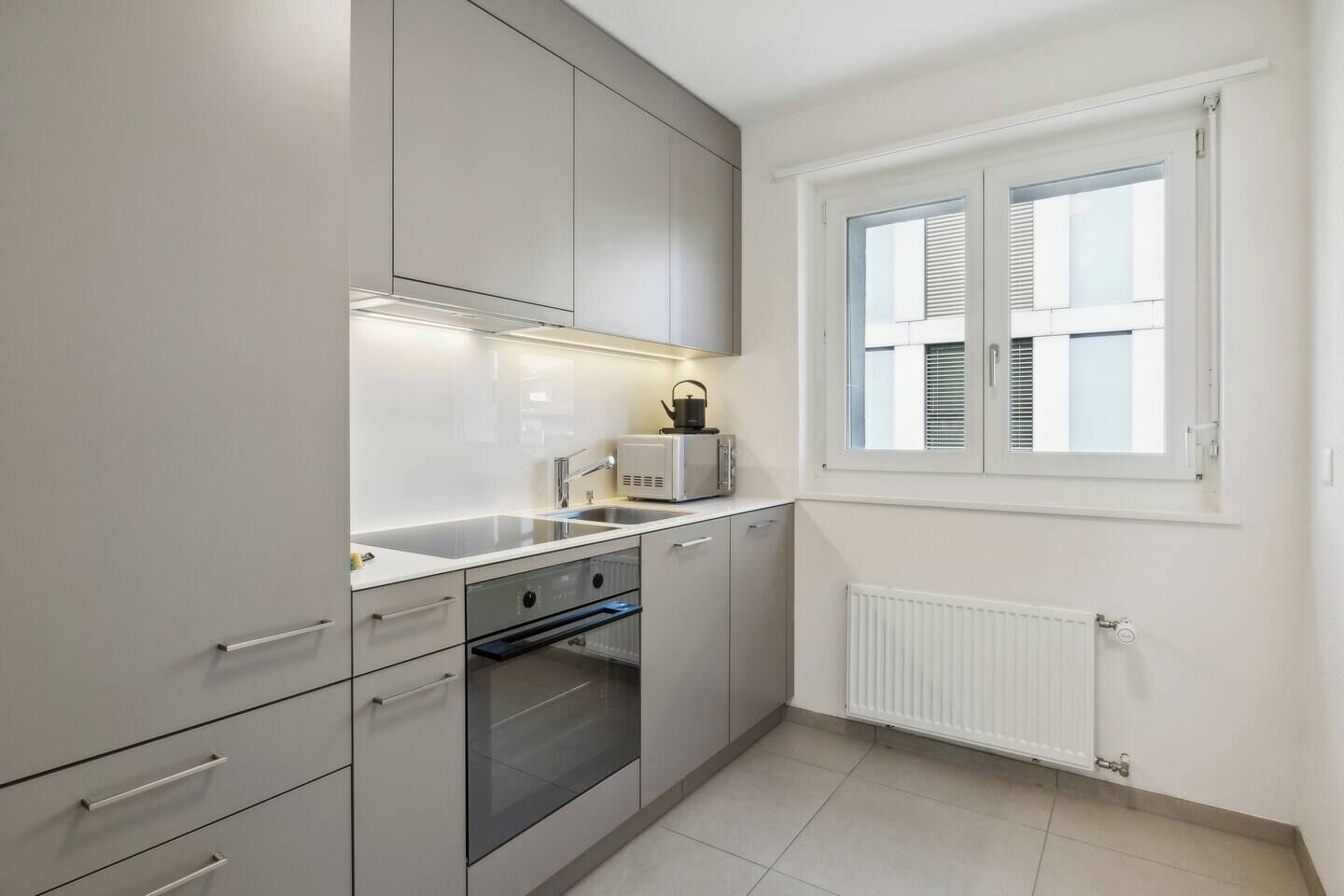 Une cuisine moderne avec des meubles gris, un four encastré, un micro-ondes et une fenêtre équipée de stores.