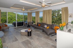 A modern living room with a sofa, coffee table, and dining area, large windows with curtains, and a ceiling fan. - Modern & Scenic Haven | Breathtaking Views - Pool (Coco)