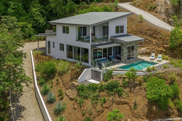Een modern huis met twee verdiepingen, met zwembad, omgeven door groen en een verharde oprit.