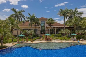 A resort with a swimming pool, palm trees, and a large building with a tiled roof. - Modern & Scenic Haven | Breathtaking Views - Pool (Coco)
