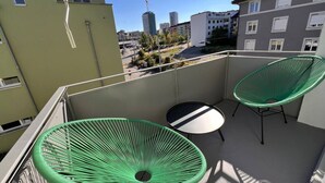 Een balkon met twee groene stoelen en een ronde tafel, met uitzicht op een stadsgezicht met gebouwen en een bouw kraan.
