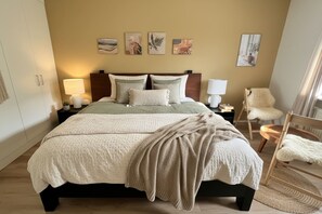 Une chambre à coucher bien aménagée, avec un grand lit, des lampes de chevet, un fauteuil moelleux et des œuvres d’art accrochées au mur.