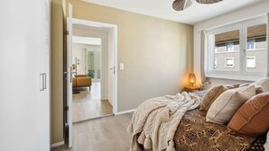 Une chambre à coucher comprenant un lit, un ventilateur de plafond, une fenêtre avec des stores et une porte donnant sur une autre pièce.