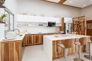 A modern kitchen with wooden cabinets, a central island with stools, a refrigerator, and a decorative wooden partition. - Luxury Ocean View Villa with Infinity Pool (Coco)