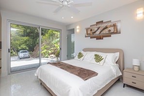 A bedroom with a bed, a nightstand, a ceiling fan, a painting, and a view of a road and greenery through a sliding door. - Luxury Ocean View Villa with Infinity Pool (Coco)