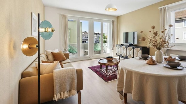 Ett modernt vardagsrum med en soffa, ett matbord med en vas med blommor och ett stort fönster med utsikt över staden.