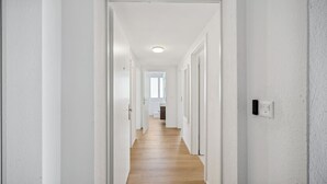 En lång, smal hall med vita väggar och trägolv, som leder till ett rum med ett fönster och en dörr längst in.