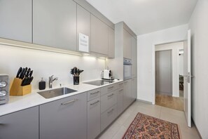 A modern kitchen with grey cabinets, a white countertop, and a built-in oven. There is a sink with a knife block and a coffee maker on the counter.