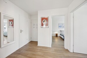 Un pasillo con paredes blancas y suelo de madera que conduce a un dormitorio con cama y mesita de noche.