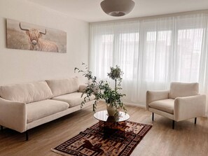Un salón con un sofá color crema, un sillón blanco, una mesa de centro con un jarrón de plantas y un cuadro grande de un toro en la pared.