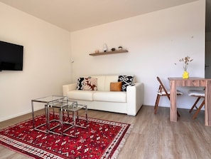 A modern living room with a white sofa, a glass coffee table, a red patterned rug, a wooden dining table, and a TV mounted on the wall. - Rooftop Suit in Zürich Oerlikon! (403) (Zürich)
