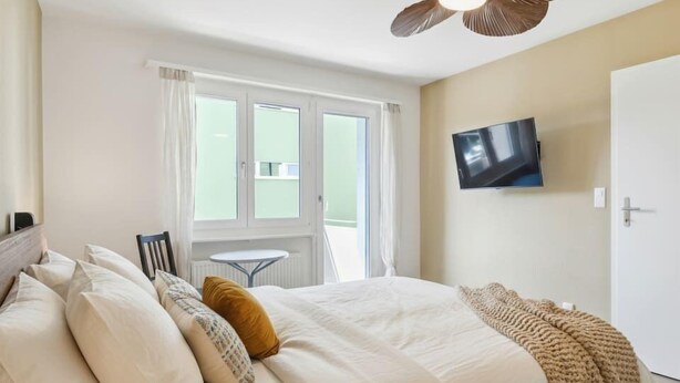 A bedroom with a bed, pillows, a TV on the wall, and a ceiling fan. - Studio w/ Balcony & Modern Amenities (202-16) (Zürich)