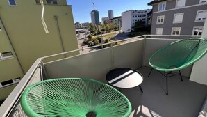 Een balkon met twee groene stoelen en een klein rond tafeltje, met uitzicht op een stadsgezicht met gebouwen en een hijskraan.