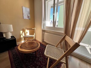 A room with a round wooden table, two wicker chairs, a radiator, a window with curtains, and a framed picture on the wall. - Elegant Studio in Zurich w/ Balcony (302-16) (Zürich)