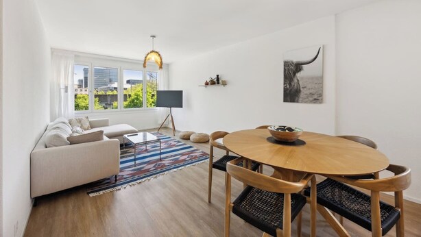 A modern living room with a sofa, a round wooden dining table with chairs, a black floor lamp, and a large window with a view of buildings and greenery. - Moderne Wohnung in Zürich Oerlikon! (201) (Zürich)
