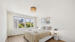 A bedroom with a large window, a bed with a patterned bedspread, a bedside table with a lamp, and two framed pictures on the wall. - Moderne Wohnung in Zürich Oerlikon! (201) (Zürich)