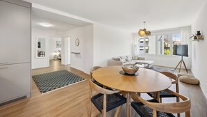 A modern living room with a round wooden table, a sofa, a TV on a tripod, and a view of the city through large windows. - Moderne Wohnung in Zürich Oerlikon! (201) (Zürich)