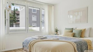 Een slaapkamer met een groot bed, een raam met gordijnen, een kamerplant en een lijst met een afbeelding aan de muur.