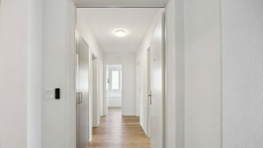 A long, narrow hallway with white walls and wooden flooring, leading to a bright room with a window.