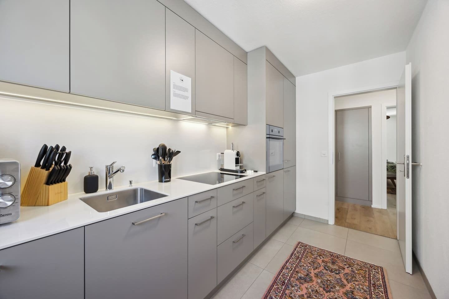 A modern kitchen with grey cabinets, a white countertop, and a built-in oven. There is a sink with a knife block and a coffee maker on the counter.