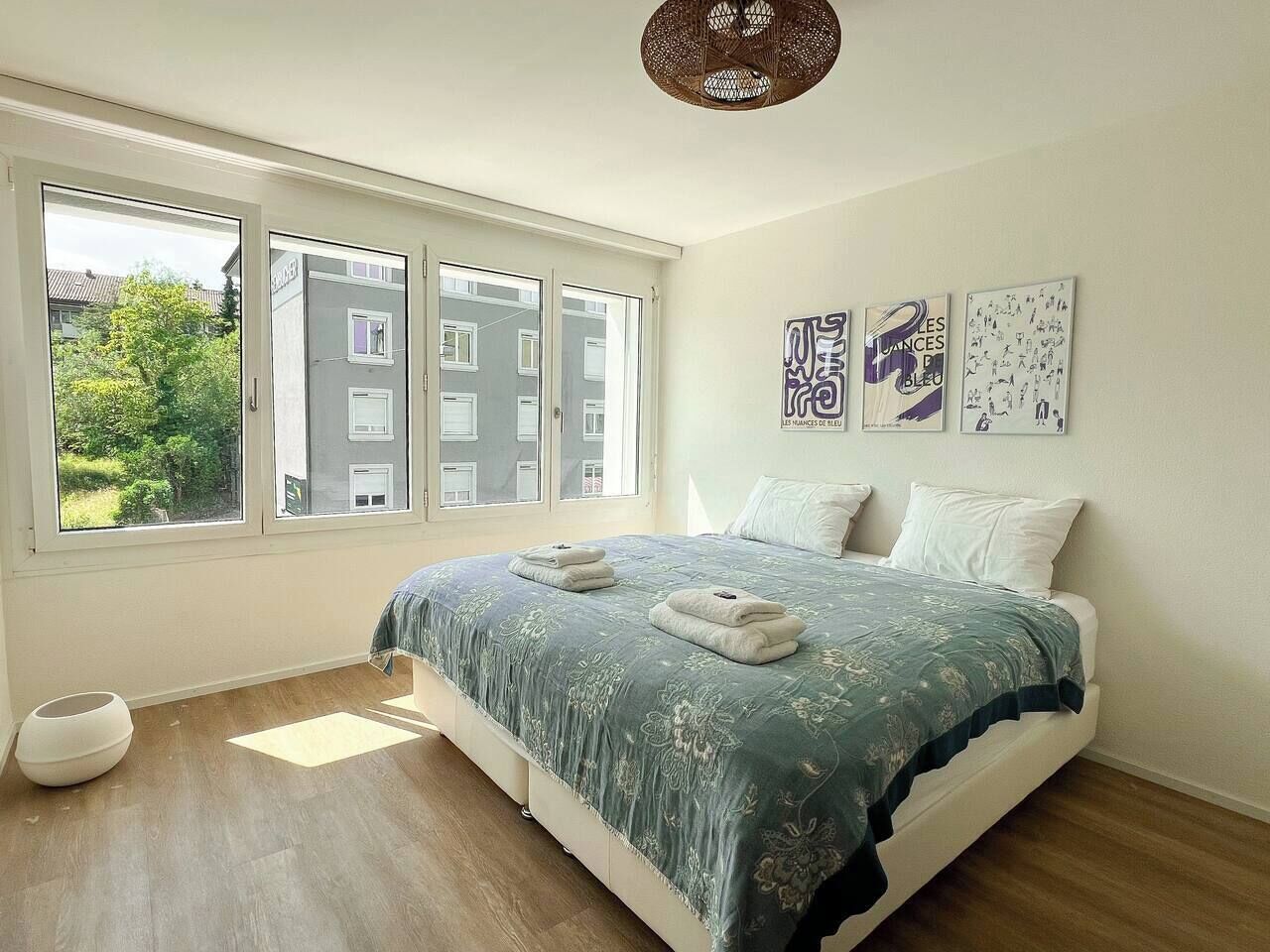 Ein Schlafzimmer mit einem großen Fenster, einem Bett mit blau-blumigem Überwurf und drei Wandbildern.