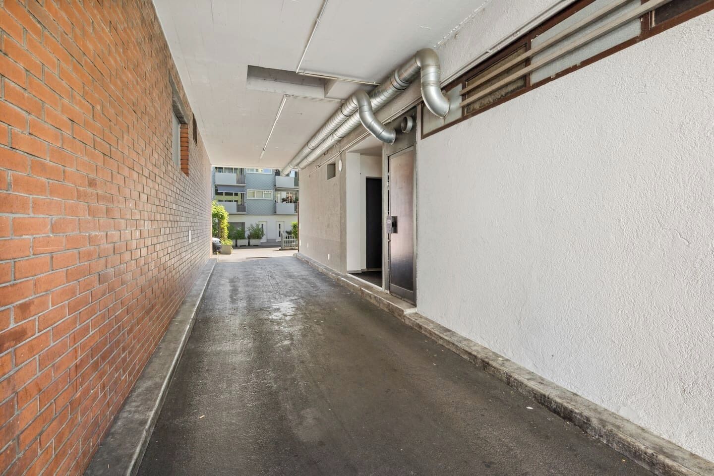 A narrow corridor with a brick wall on one side and a white wall on the other, leading to a door with a metal handle.