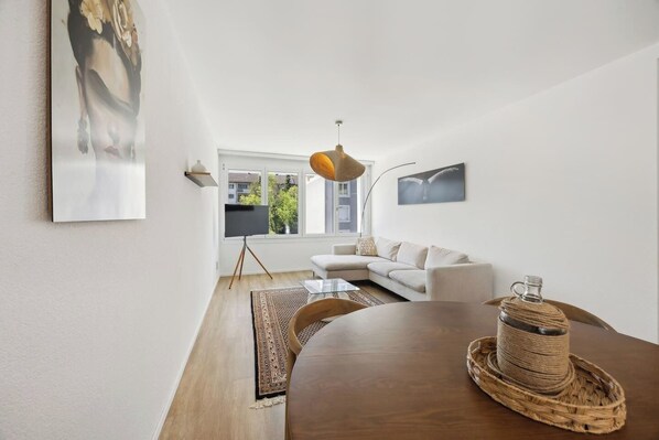 Un salon moderne avec une table à manger en bois, un canapé blanc et une grande fenêtre donnant sur des immeubles.