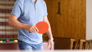 Iemand houdt een tafeltennisracket vast in een ruimte met een pooltafel, houten wanden en een biljartopstelling.