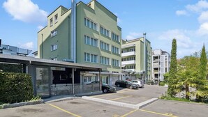 Un immeuble à plusieurs étages, doté d’une façade verdoyante, d’une entrée abritée et d’un espace de stationnement où sont garées des voitures.