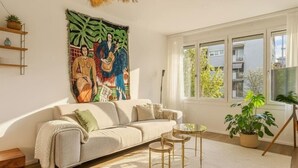 Un salon moderne avec un canapé beige, un grand tableau abstrait, une table basse en verre et une vue sur les bâtiments à l’extérieur.